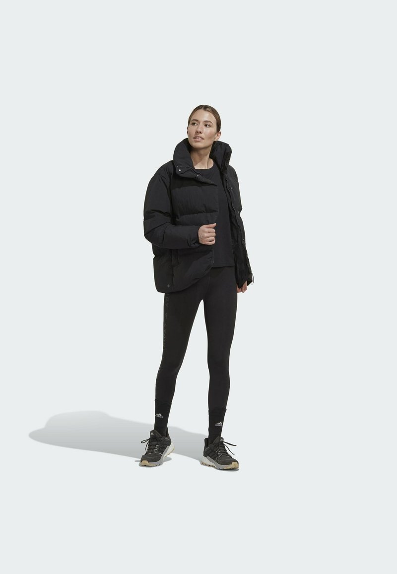 Schwarze Pufferjacke mit hohem Kragen, Reißverschluss und Druckknöpfen, kombiniert mit schwarzen Leggings und grauen Sportschuhen. Texturiertes Material.