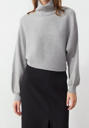 Femme portant un pull à col roulé côtelé gris clair avec des manches ballon et une jupe noire taille haute avec une fente à l'avant.