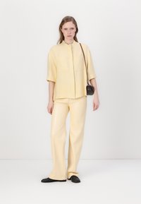 Ljusgul kortärmad skjorta med två bröstfickor, i kombination med matchande vida byxor. Svarta platta skor fulländar outfiten.