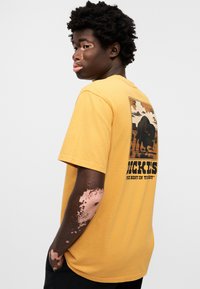 Κίτρινη βαμβακερή μπλούζα με γραφικό σχέδιο βίσονου στην πλάτη, με μαύρο κείμενο και ορθογώνιο πλαίσιο. Κοντά μανίκια και άνετη εφαρμογή.