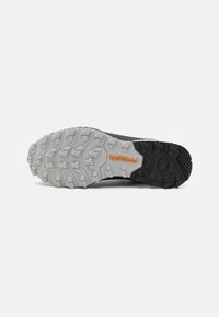 Suola di scarpa da trail running con un aggressivo motivo a scanalature in gomma, sfumatura da grigio a nero, con un dettaglio del logo arancione.