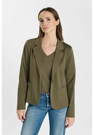 Vrouw met lang bruin haar die een olijfgroene blazer en bijpassend V-hals shirt draagt, gecombineerd met lichtblauwe spijkerbroek tegen een effen witte achtergrond.