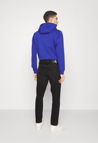 Blauwe sweatshirt met capuchon, gecombineerd met zwarte jeans. De outfit heeft een strak ontwerp, textuurstof en een subtiele logo op de achterkant.