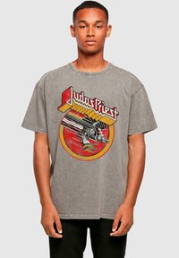 Graues, übergroßes T-Shirt mit einem auffälligen roten und gelben Grafikmotiv einer Hand und dem Text "Judas Priest", in einer weichen, abgenutzten Textur.