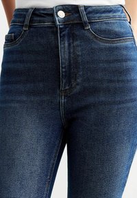 Jeans de mezclilla azul de cintura alta con una textura suave, diseño de cinco bolsillos y un botón plateado en la cinturilla. Apariencia clásica de lavado oscuro.