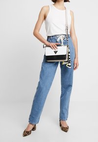 Donna in top senza maniche bianco, jeans blu a vita alta, tacchi con stampa leopardo, che tiene in mano una borsa designer bianca e nera con un accessorio sciarpa.