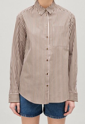 Osoba ubrana w brązowo-białą, pionowo prążkowaną koszulę z długim rękawem i guzikiem, połączoną z niebieskimi krótkimi spodniami jeansowymi.