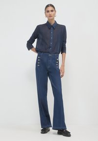 Camicia trasparente blu navy con bottoni e righe; pantaloni in denim a vita alta e gamba larga con dettagli in bottone dorato ai lati; abbinati a scarpe nere.
