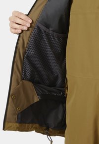 Main tenant une veste marron ouverte montrant une poche intérieure en maille noire et un cordon de serrage réglable à l'ourlet inférieur.