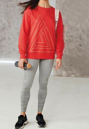 Felpa rossa con design geometrico dorato, leggings grigi con motivo camouflage e scarpe da ginnastica nere. Bottiglia d'acqua tenuta in mano.