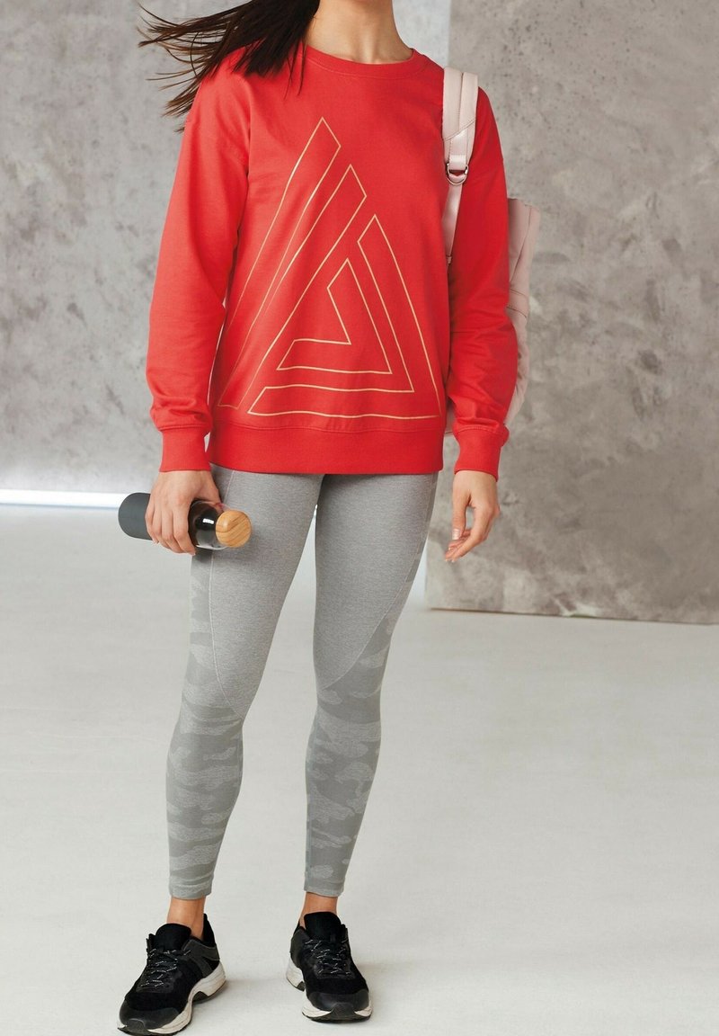 Rode sweatshirt met gouden geometrisch ontwerp, grijze legging met camouflagepatroon en zwarte sportschoenen. Waterfles in de hand gehouden.