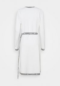 Robe blanche à manches longues avec un col rond, une ceinture imprimée du logo et des poignets. Texture lisse avec une coupe ajustée et une longueur jusqu'au genou.
