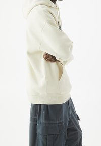 Hoodie de couleur crème avec une poche frontale et des poignets côtelés, associé à un pantalon cargo sombre avec plusieurs poches et des détails de couture.