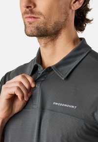 Grå polo tröja med ett texturerat mönster, med krage och knäppning. "SWEDEMOUNT" logotyp i vitt på bröstet.