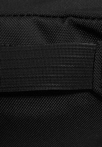 Tessuto nero con texture e motivo a griglia, dotato di una tracolla piatta nella parte superiore. La superficie sembra resistente e robusta.