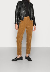 Femme portant un pantalon en velours côtelé marron, une veste en cuir noire, un col roulé noir et des mocassins noirs, debout sur un sol gris contre un mur blanc.