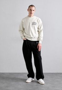 Felpa cremosa con logo "Aries" grigio, abbinata a pantaloni neri e sneakers bianche. Lo sfondo è di un grigio chiaro uniforme.