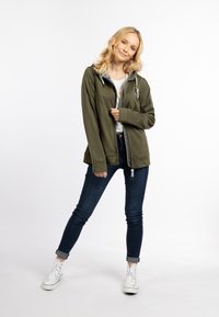 Giacca verde oliva con zip e cappuccio, due tasche frontali e coulisse regolabili. Abbinata a jeans scuri e sneakers bianche.