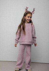 Młoda dziewczynka uśmiechająca się, ubrana w różowy sweter z dzianiny oraz spodnie, z pluszowym diademem w kształcie renifera na głowie, na jednolitym tle.