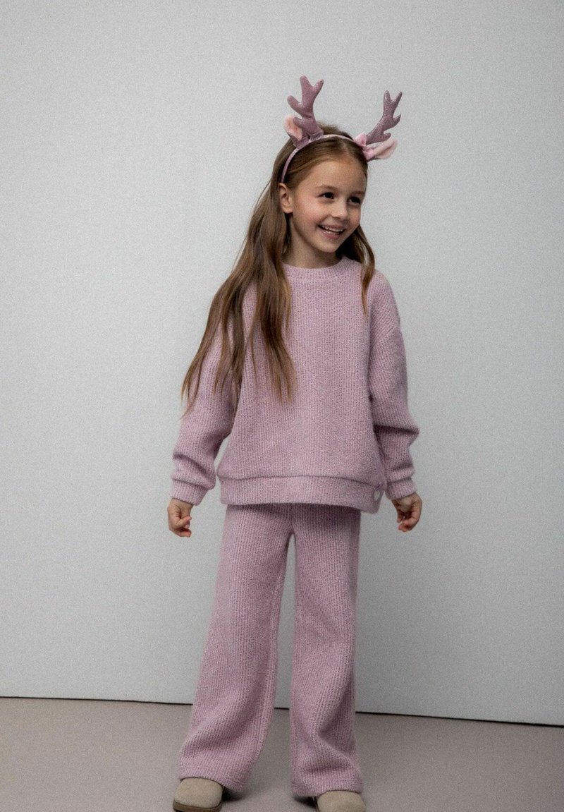 Młoda dziewczynka uśmiechająca się, ubrana w różowy sweter z dzianiny oraz spodnie, z pluszowym diademem w kształcie renifera na głowie, na jednolitym tle.