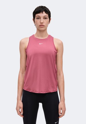 Femme aux cheveux courts et foncés portant un débardeur rose Nike sans manches et des leggings noirs avec des logos Nike blancs, debout devant un fond uni.