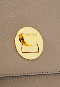 Borsa a tracolla in pelle marrone chiaro con finitura texture, dotata di un pattino rotondo color oro inciso con "Coccinelle". Dettagli di cucitura minimi visibili.