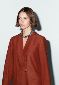 Blazer color ruggine con finitura texturizzata, caratterizzato da un profondo scollo a V e un singolo bottone. Collana dichiarativa bianca e nera che aggiunge contrasto.