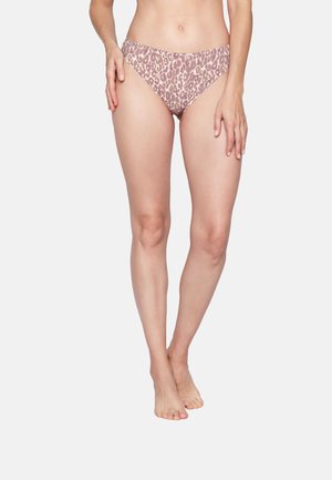 Frau steht barfuß in hoch geschnittenen Leopardenmuster-Bikinihosen, Arme entspannt an den Seiten, vor einfachem hellem Hintergrund.