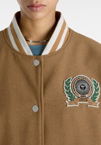 Brun uld varsity jakke med stribet krave, prydet med et cirkulært broderet logo og trykknaplukning foran. Glat tekstur.
