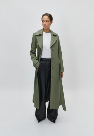 Γυναίκα που φοράει μακρύ παλτό ελιάς, λευκό πουκάμισο, μαύρο παντελόνι φαρδύ και μαύρα μυτερά παπούτσια, στέκεται με το ένα χέρι στην τσέπη.