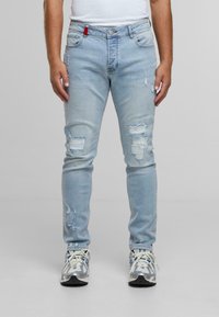 Light blue denim jeans with a slim fit, featuring multiple distressed areas and a red accent on the waistband. Paired with gray sneakers.