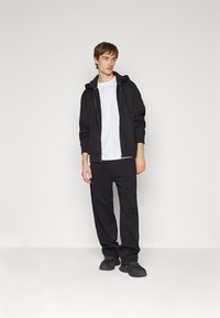 Sudadera negra con cierre frontal y bolsillo delantero, combinada con pantalones negros holgados y zapatos negros, sobre un fondo gris liso.