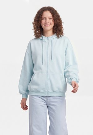 Tânăr cu păr creț purtând un hanorac bleu deschis cu fermoar și pantaloni deschiși la culoare, zâmbind pe fundal alb simplu.