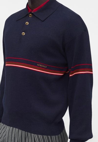 Homme portant un pull polo bleu marine avec une bande rouge, marron et blanche sur la poitrine et des boutons dorés.