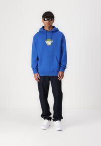 Blue cotton hoodie with a front pocket and a yellow-green graphic logo. Paired with dark denim jeans and white sneakers.