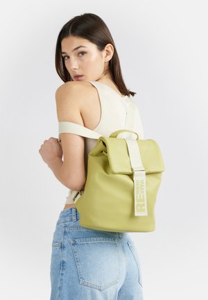 Giovane donna con canottiera leggera e jeans, che porta uno zaino chartreuse con una tracolla bianca su una spalla.