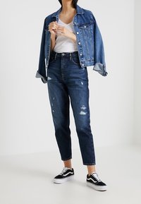 Jeansjacka, ljusblå, avslappnad passform, med knappar och fickor; buren över en vit t-shirt. Mörkblå slitna jeans med smalnande ben. Svarta sneakers.