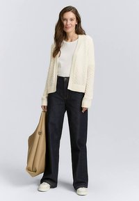 Krém színű kötött kardigán bordázott mandzsettákkal és szegéllyel, fehér felső és sötét, széles szárú farmernadrág fölött viselve, világosbarna válltáskával és fehér cipővel párosítva.