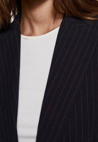 Námořnický blazer s jemným proužkem, se strukturovaným límcem a klopami, spárovaný s přiléhavým bílým ribbed topem. Hladká textilie, moderní design.