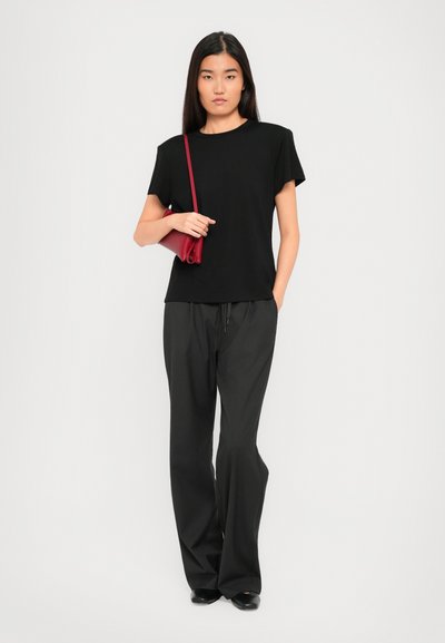 Woman wearing black t-shirt and wide-leg pants, holding a red shoulder bag, standing against a plain white background.