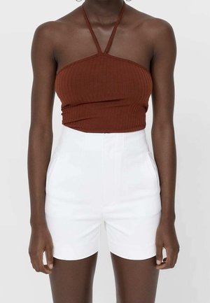 Mørkhudet person iført en brun ærmeløs halter crop top og højtaljede hvide shorts mod en ensfarvet baggrund.