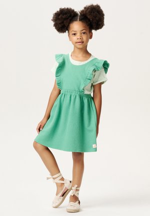 Grünes, strukturiertes Kleid mit Rüschensleeven, getragen über einem gestreiften Shirt. Geflochtene beige Sandalen mit Schleifen am Knöchel. Kleines Logo auf dem Kleid.
