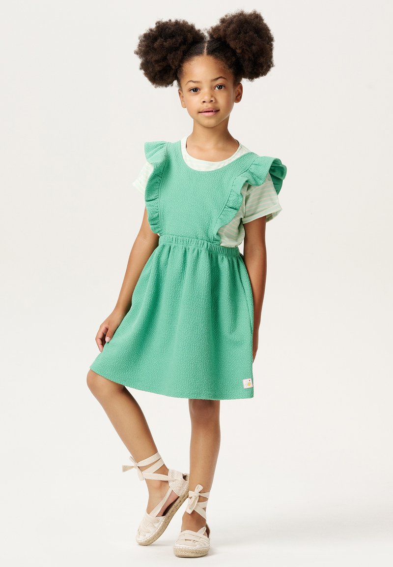 Zielona sukienka o teksturze z frędzlami na rękawach, noszona na paskowaną koszulę. Plecione beżowe sandały z kokardkami przy kostce. Małe logo na sukience.