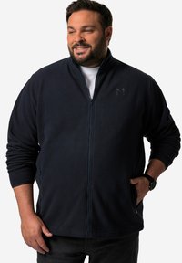 Giacca in fleecia blu navy con colletto alto, zip frontale e tasche laterali. Presenta un sottile logo "M" sul petto e polsini a costine.