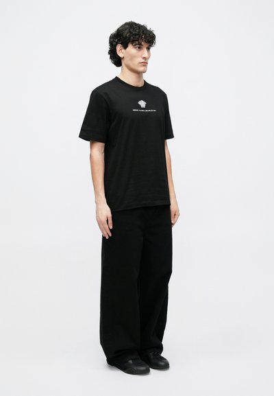 Μαύρο βαμβακερό μπλουζάκι με κοντά μανίκια που έχει λευκό γραφικό κείμενο στο μπροστινό μέρος, συνδυασμένο με φαρδιά μαύρη παντελόνια και μαύρα παπούτσια.