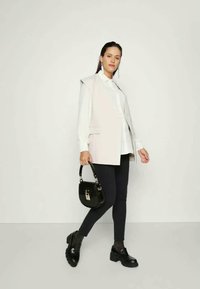 Blazer sem mangas cinza claro sobre uma camisa branca, combinado com calças skinny escuras e sapatos pretos chunky; segurando uma pequena mala preta.