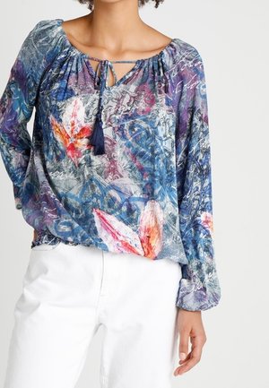 Kvinde iført en langærmet, flerfarvet blomsterbluse med en snøre med frynser i halsen, kombineret med hvide bukser.