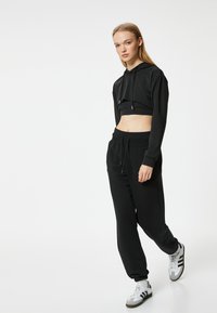 Afgesneden zwarte hoodie met een drawstring en lange mouwen, gecombineerd met losse zwarte joggingsbroeken. Het model draagt witte en grijze sneakers.