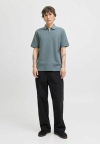 Lichtgroen polo-shirt met een ritskraag, korte mouwen en een gestructureerde stof, gecombineerd met zwarte wijde broek en donkere schoenen.