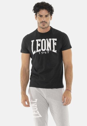 Homme aux cheveux bouclés foncés portant un t-shirt noir avec "LEONE 1947" et un pantalon de jogging gris clair avec "LEONE" imprimé sur la jambe.